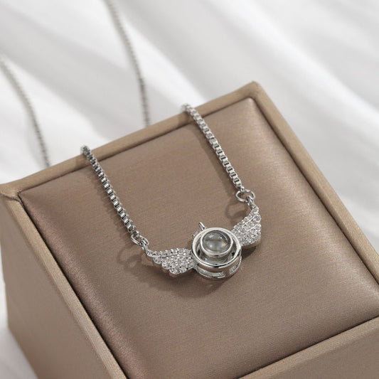 May include: A silver necklace with a clear crystal pendant in the shape of angel wings. The pendant has a small, round, silver-colored projection inside. The necklace is on a brown velvet surface.