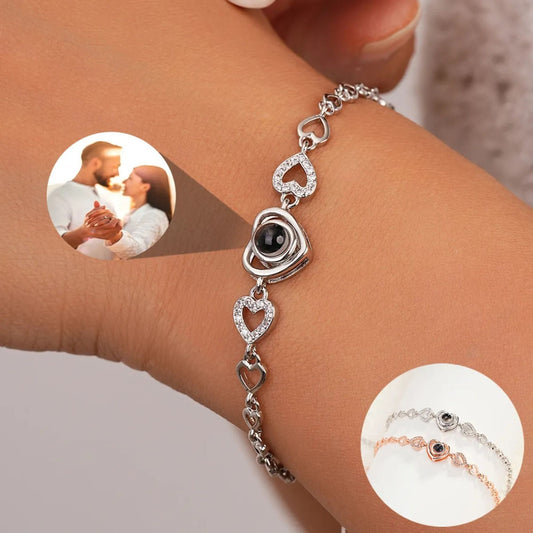 May include: A silver bracelet with a heart-shaped pendant that projects an image when held up to a light source. The bracelet is adorned with small, clear cubic zirconia stones. The pendant is surrounded by a ring of smaller hearts. The bracelet is shown on a person&#39;s wrist.