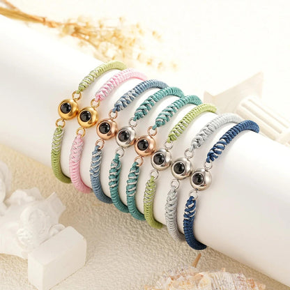 May include: A set of seven colorful friendship bracelets with a silver and black eye charm. The bracelets are made of braided string and come in colors like green, pink, blue, and gray.