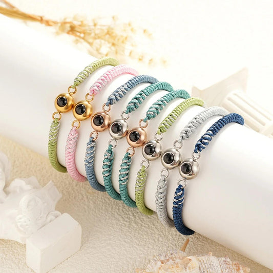 May include: A set of seven colorful friendship bracelets with a silver and black eye charm. The bracelets are made of braided string and come in colors like green, pink, blue, and gray.